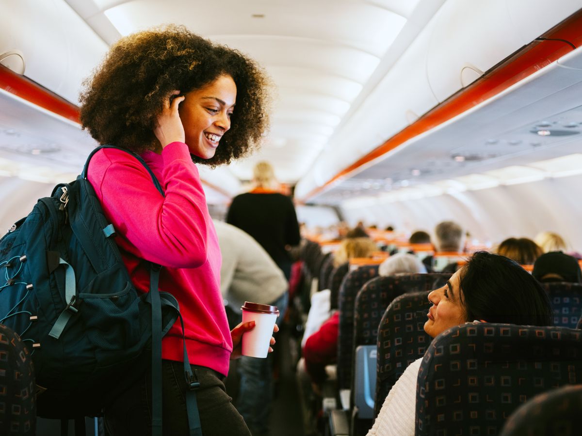 Passenger in airplane aisle conversing with friend in seat