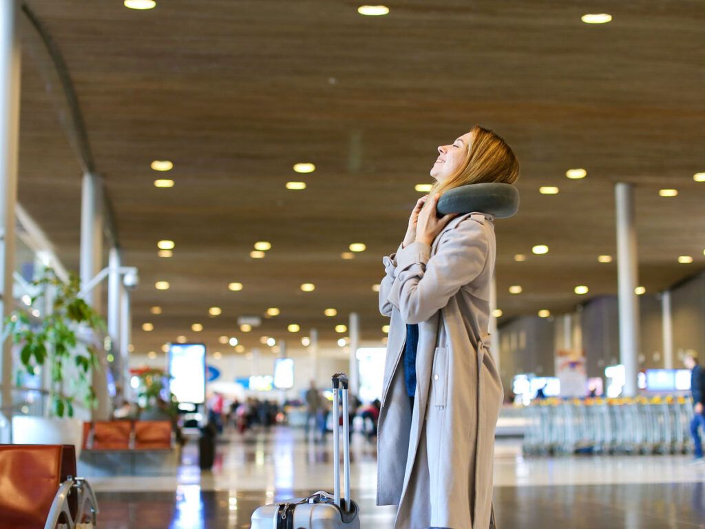 Passenger standing in airport terminal with suitcase and neck pillow